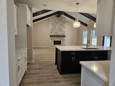 Open-concept living room featuring exposed wooden beams, a stone fireplace, and a modern kitchen with dark cabinets.