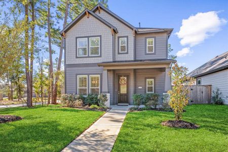 This is a two-story home with a gray exterior and white trim, featuring large windows, a covered entryway, and a well-maintained lawn with young trees. The property is flanked by tall pine trees and has a welcoming, suburban feel.