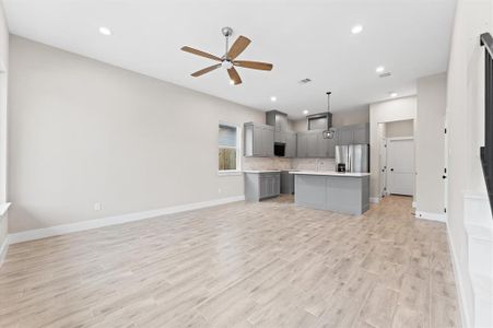 This spacious open-concept living area features modern gray cabinetry, a sleek kitchen island, stainless steel appliances, and elegant wood-look tile flooring. The room is bright with recessed lighting and a stylish ceiling fan, creating a welcoming atmosphere.