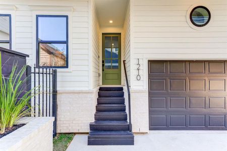 This photo showcases a modern stairway entrance with a stylish green door, black accented double  paned windows, adjacent to a  matching garage door. The exterior features Hardie Plank siding and neutral bricks, and a small landscaped area  with shrubs.