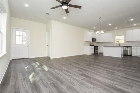 A spacious empty room with gray flooring, white kitchen cabinets, modern appliances, and a ceiling fan, illuminated by natural light.