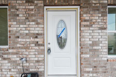 A white front door with oval glass, framed by a brick wall, and a lawnmower cable plugged into an outdoor outlet to the left.