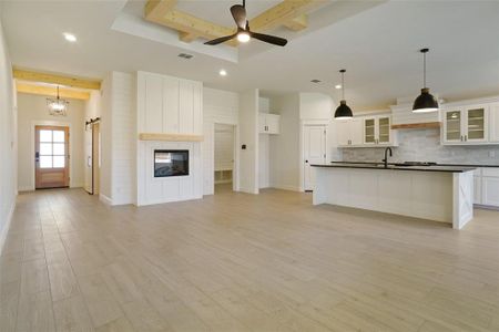 Kitchen featuring a large fireplace, white cabinetry, light wood-type flooring, an island with sink, and beamed ceiling