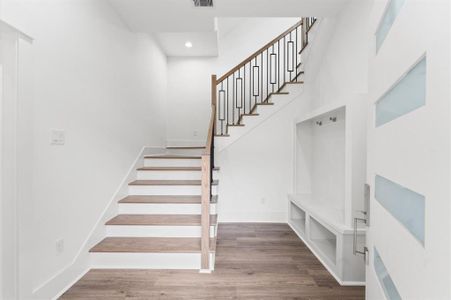 This photo showcases a modern entryway with a stylish staircase featuring wooden steps and a sleek metal railing. The space is bright with white walls, a contemporary door with frosted glass inserts, and a built-in bench with storage. The flooring is a rich, warm wood, adding elegance to the area.