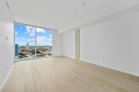 Living room with large, floor-to-ceiling glass doors leading to a balcony. The view overlooks a cityscape with prominent buildings, including the University of Texas. The room has light-colored wood flooring and a clean, modern aesthetic with a minimalist design.