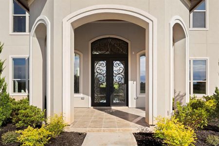 Elegant front entryway to a modern home, showcasing an arched doorway with a decorative wrought iron door, flanked by tall windows and completed with neatly landscaped bushes.