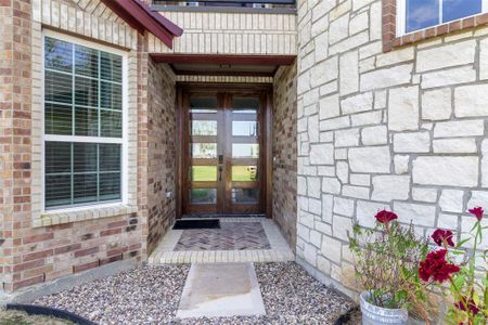 This inviting entryway features a wooden double front door framed by brick and stone accents. The walkway is lined with decorative gravel and leads to a cozy, covered porch area. A large window allows natural light to brighten the space, and a touch of greenery adds charm.