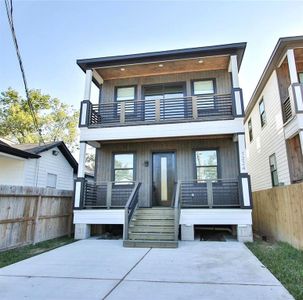 This modern two-story home features a sleek wood and black exterior, with two balconies and a welcoming entryway. The property includes a paved driveway and is surrounded by a wooden fence, offering privacy and a contemporary feel.