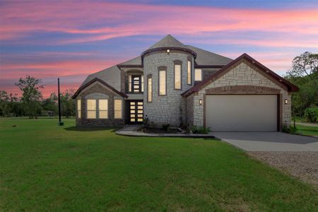 This photo showcases a spacious two-story stone house with a unique turret feature and a large, well-maintained lawn. The home has a two-car garage and is set against a beautiful sunset sky, providing a serene and inviting atmosphere.