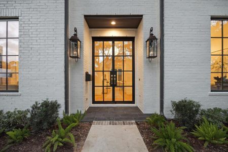 The home showcases a modern white-painted brick facade with tall black steel and glass double front doors. Gas lanterns on each side, w/ a concrete pathway lined with manicured shrubs leads to the black slate-covered entryway