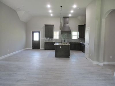 Kitchen with custom range hood, light stone counters, a kitchen island with sink, decorative backsplash, and vaulted ceiling