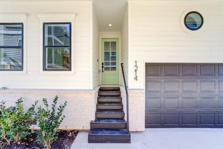 This photo showcases a modern home entrance with a stylish light green door, black accented double paned windows, adjacent to a matching garage door. The exterior features clean white Hardie Plank siding and a small landscaped area with shrubs.