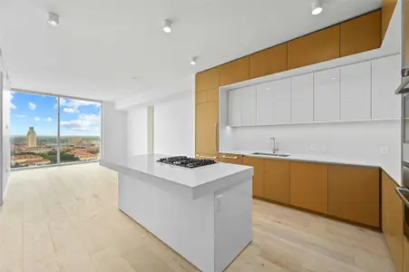 A modern, open-concept kitchen, dining and living area. The kitchen features a sleek, minimalist design with a large island at its center, topped with white-colored stone countertop. The island includes a built-in gas cooktop and stainless steel oven.