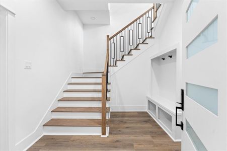 This photo shows a modern, bright entryway with a wooden staircase featuring sleek metal railings. The space includes built-in storage and hooks, perfect for organization, and has a stylish white door with frosted glass panels. The overall design is clean and contemporary.