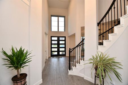 Spacious and elegant entryway with high ceilings, modern dark front door, and a curved staircase with black iron railing. Bright and welcoming, with light wood flooring and potted plants adding a touch of greenery.