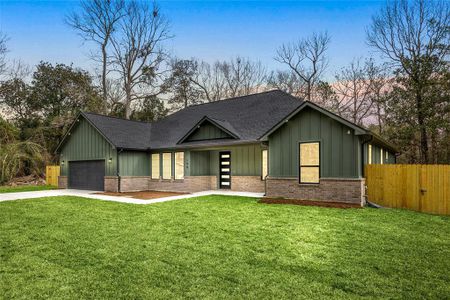 This photo features a modern, single-story home with a green exterior and dark roof. It has a two-car garage, large front windows, and a spacious, well-maintained lawn, bordered by a wooden fence, with a backdrop of mature trees providing a serene setting.