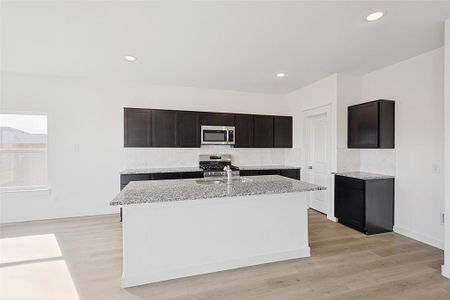 Kitchen with light stone counters, light hardwood / wood-style flooring, a kitchen island with sink, dark brown cabinets, and appliances with stainless steel finishes