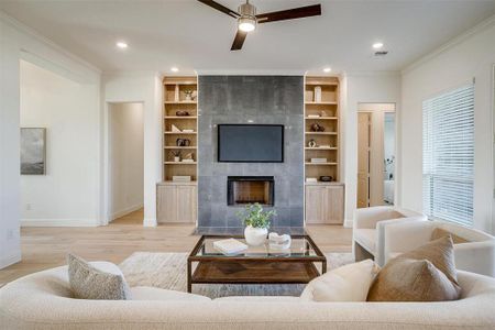 Living room with light wood-type flooring, ornamental molding, ceiling fan, built in cabinetry, and a tiled fireplace