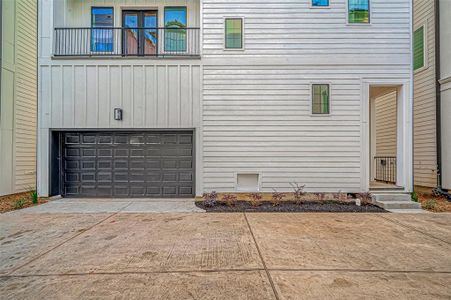 This photo shows a modern townhouse exterior with clean white siding, a dark garage door, and a small landscaped area. The entrance features a covered walkway with steps leading up. The design is sleek and contemporary.