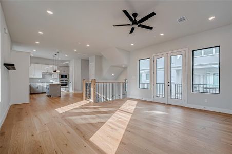 A spacious, open-concept living area with hardwood floors, abundant natural light, and a modern kitchen featuring white cabinetry and a central island. The room includes recessed lighting, a ceiling fan, and access to a balcony through large double doors, offering a bright and inviting atmosphere.