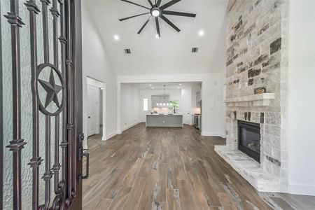 This inviting home features a spacious open-concept living area with a high ceiling and modern ceiling fan. The room boasts elegant wood-look flooring and a striking stone fireplace. The kitchen, visible in the background, is well-equipped and stylish, perfect for entertaining.