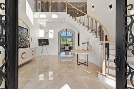 Grand foyer with high ceilings, polished floor tiles, and an elegant curved staircase with wrought-iron railings. The space is well-lit, with natural light streaming in through large windows, and features a neutral color palette that offers a sense of luxury and spaciousness.