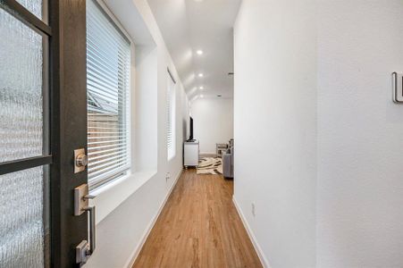 You are greeted through a bright, narrow entryway with wood flooring, leading into a cozy living area. Large windows on the left provide natural light, creating a welcoming atmosphere.