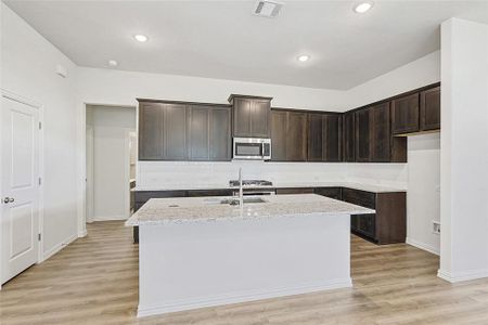 Kitchen with light stone countertops, dark brown cabinets, a center island with sink, and light hardwood / wood-style flooring