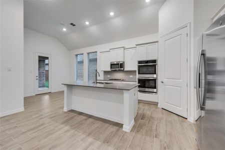 Kitchen featuring light wood-type flooring, appliances with stainless steel finishes, an island with sink, backsplash, and vaulted ceiling