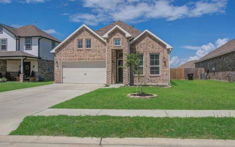 This is a modern single-story brick home featuring a two-car garage, a well-maintained lawn, and a simple landscaping design with a young tree in the front yard. The house has a prominent gable roof and a welcoming entrance.