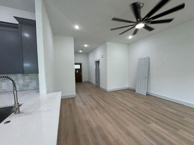 This is a modern, open-plan living space with sleek dark cabinetry in the kitchen, light wood-look flooring, and a contemporary ceiling fan. The room is well-lit and appears to lead to additional rooms, providing a sense of spaciousness.