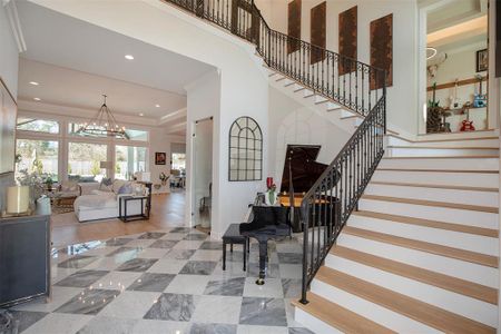 This elegant home features a spacious, open living area with large windows providing ample natural light. A grand staircase with ornate railings leads to the upper level. The entryway includes polished marble flooring and a stylish piano, enhancing the sophisticated ambiance.