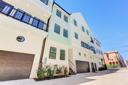 This photo shows a modern multi-story single-family home with a clean, white exterior and dark garage door. The building features a second story balcony with sleek railings and well-maintained landscaping. It is situated in the Heights on a private driveway.