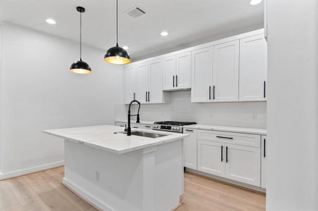 Modern kitchen with a sleek design, featuring white cabinetry, black hardware, and a central island with a sink. The space is well-lit with pendant lights and recessed lighting, complemented by light wood flooring.