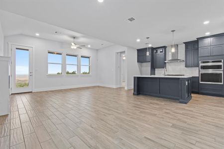 Kitchen featuring stainless steel double oven, wall chimney exhaust hood, hanging light fixtures, ceiling fan, and a center island with sink