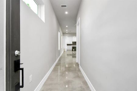 This is a modern, well-lit hallway with polished concrete floors, leading into an open-plan area with a glimpse of the kitchen in the distance. The space feels clean and spacious, featuring high ceilings and recessed lighting.