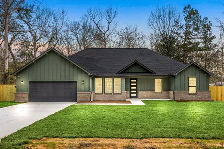 Modern single-story home with a dark green exterior and brick accents, featuring a spacious front lawn. It includes a two-car garage and a stylish front entrance with large windows. The property is bordered by a wooden privacy fence, surrounded by mature trees.