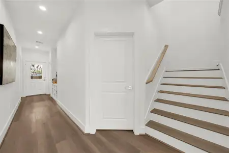 Well-lit hallway with wooden flooring, a staircase, and a white door. The open space is bright, leading to Two downstairs bedrooms and a patio area.