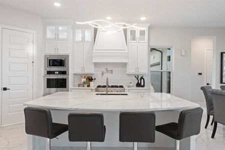 Kitchen featuring light stone counters, decorative backsplash, hanging light fixtures, an island with sink, and white cabinetry