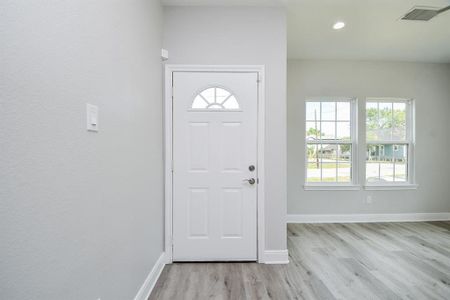 This photo shows a bright, modern entryway with a white front door featuring an arched window, complemented by a large double window that allows for ample natural light. The space has light grey walls and wood-look flooring, creating a clean and welcoming atmosphere.