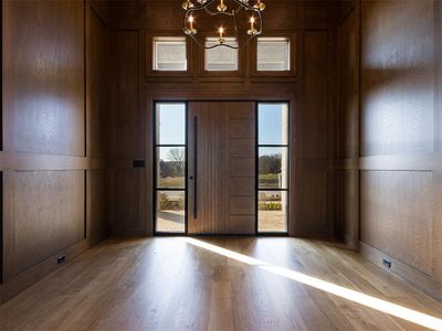 Light and bright foyer entrance with wood door and flooring, a designer chandelier, and surrounded in white oak paneling.