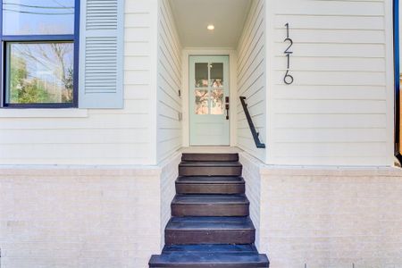 This photo showcases a modern fenced Victorian home entrance with a stylish door, black accented double paned windows, adjacent to a matching garage door. The exterior features clean white Hardie Plank siding neutral brick, and a small landscaped area with shrubs.