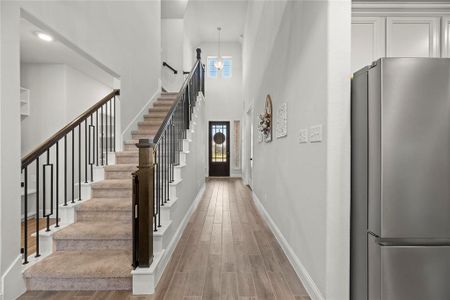 This is a modern entryway featuring a carpeted staircase with a wooden handrail and iron balusters, high ceilings, and wood-look flooring. A stainless steel refrigerator is to the right, indicating an open layout with the kitchen adjacent to the entrance. The space is well-lit with natural light coming from the door with glass elements.