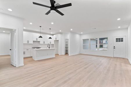 This open-concept living space features a modern kitchen with white cabinets, an island, and pendant lighting. The area is bright with recessed lighting and large windows, complemented by light wood flooring and a ceiling fan. The entry door adds a charming touch.