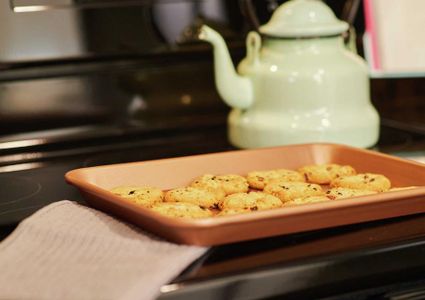 Staged photo stovetop detail with cookies in tray, kettle on black electric cooktop.