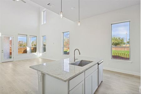 Kitchen featuring sink, dishwasher, light stone counters, decorative light fixtures, and a center island with sink