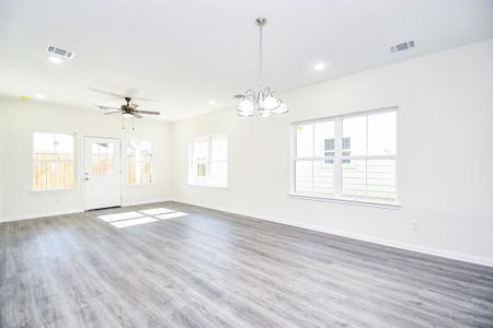 Bright, empty room with wood flooring, white walls, several windows, ceiling fan, and a chandelier; appears to be a modern living space.