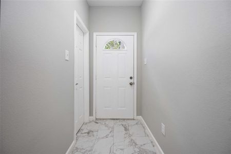 This photo shows a bright, modern entryway with light gray walls and marble-patterned tile flooring. The white front door features an arched window, adding a touch of elegance to the space.