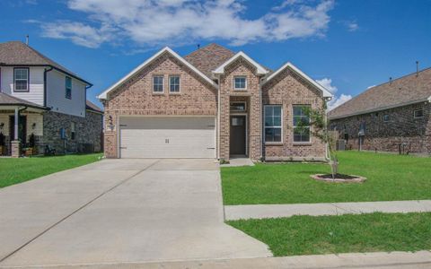 This is a single-story, brick-front home featuring a two-car garage, a well-manicured lawn, and a small tree in the front yard. The house has a pitched roof and a prominent entryway. It is situated in a suburban neighborhood with similar properties adjacent to it.