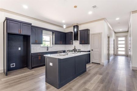 Kitchen with light wood-type flooring, an island with sink, decorative light fixtures, and wall chimney range hood